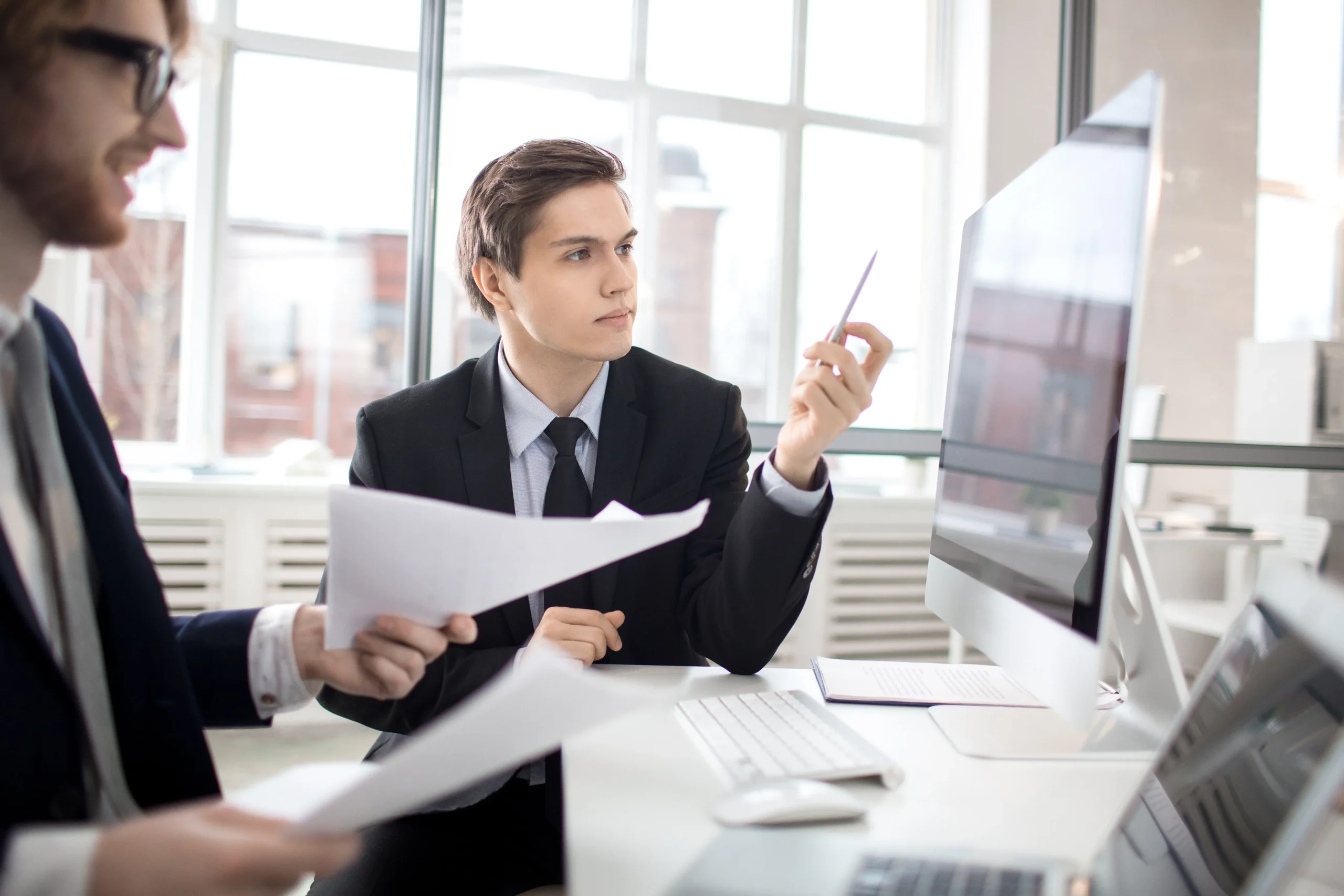 Two business professionals discussing data reports at a desk with a desktop computer, showing how a Regular SAM Screening should be part of a corporate compliance program.