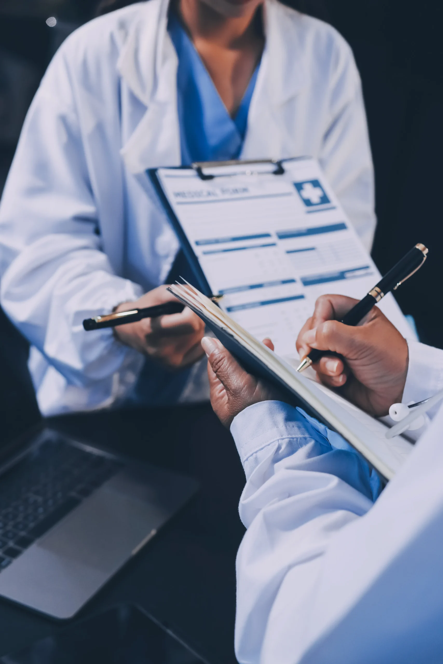Healthcare staff writing notes on clipboards to manage a compliance workflow near a laptop.