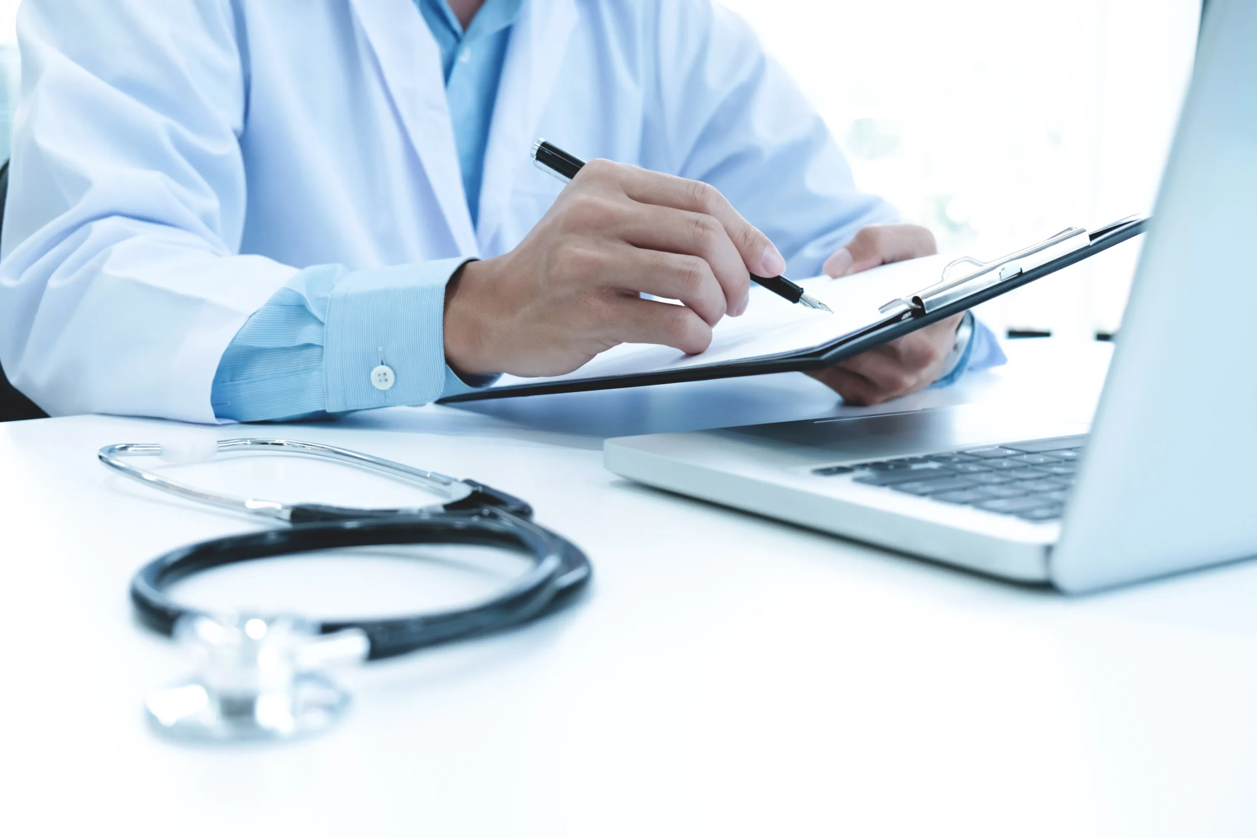 Doctor taking notes on a clipboard beside a laptop running a healthcare compliance management system.